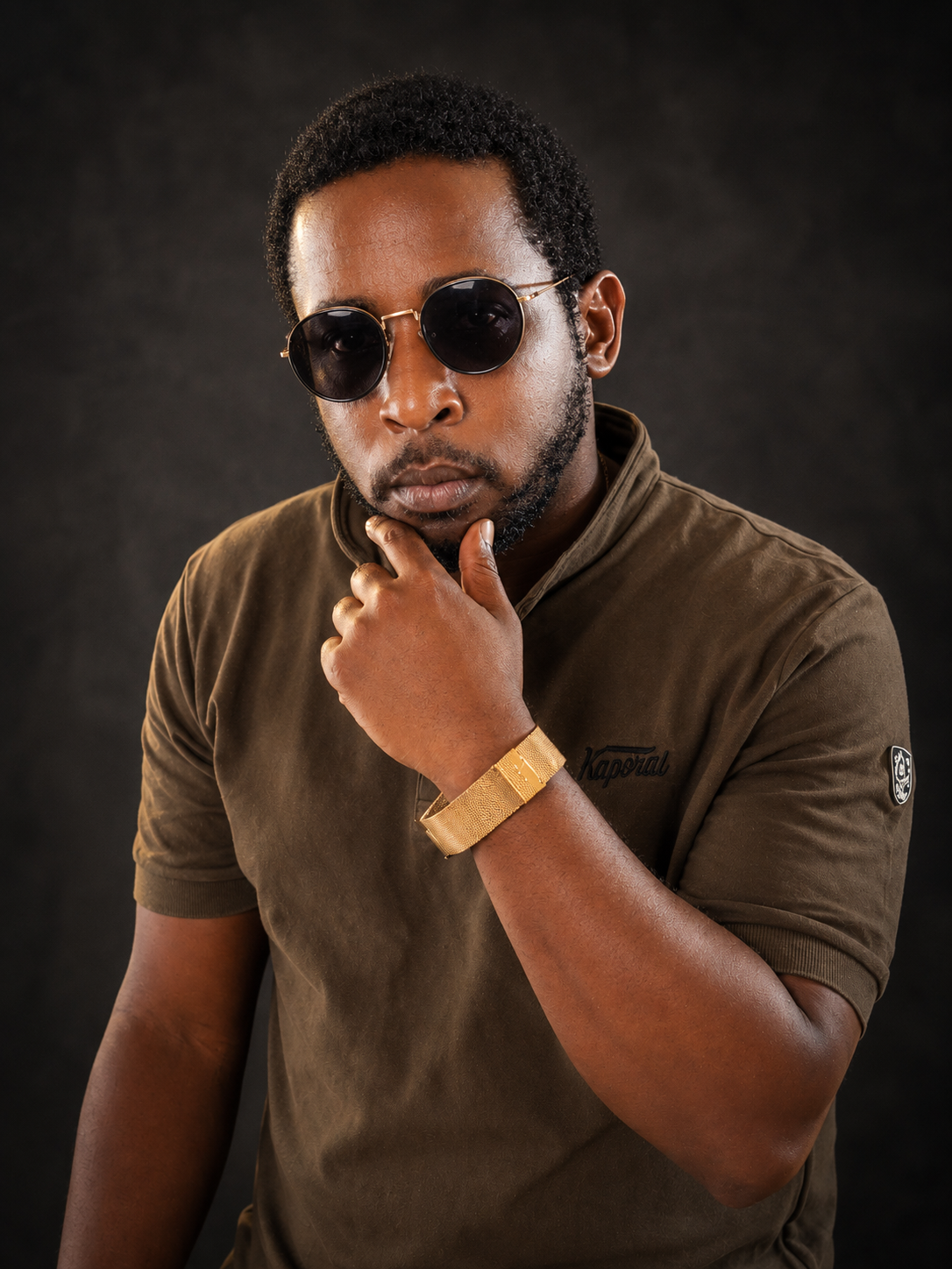 A stylish Ochomane wearing round sunglasses and a gold bracelet posing against a dark background.